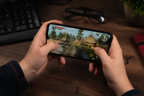 Ręce trzymają smartfon, na którym widać fragment gry z widokiem na wioskę. To dowód, że najlepsze gry na telefon to teraz norma.